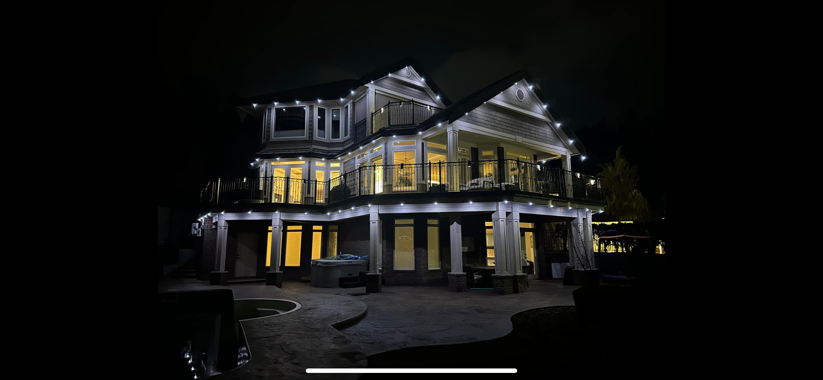Permanent bright-white accent lighting outlining a large two-story home, deck and railings at night