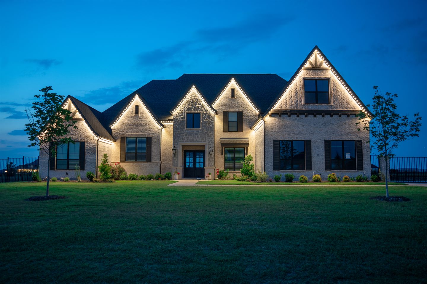 A stone-fronted home at dusk with warm white permanent lighting tracing every roofline, gable and peak.